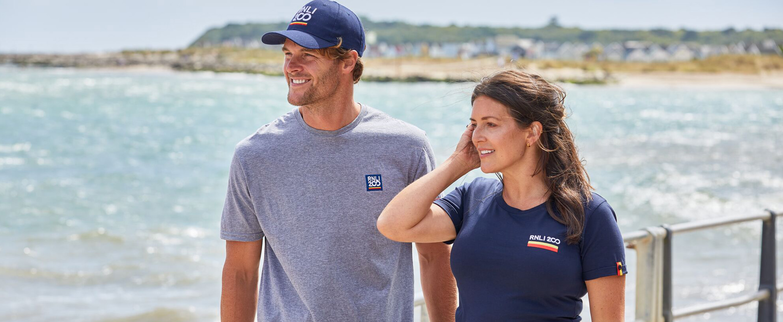 A woman and a man, wearing RNLI 200 T-shirts, stand on a promenade in front of the sea