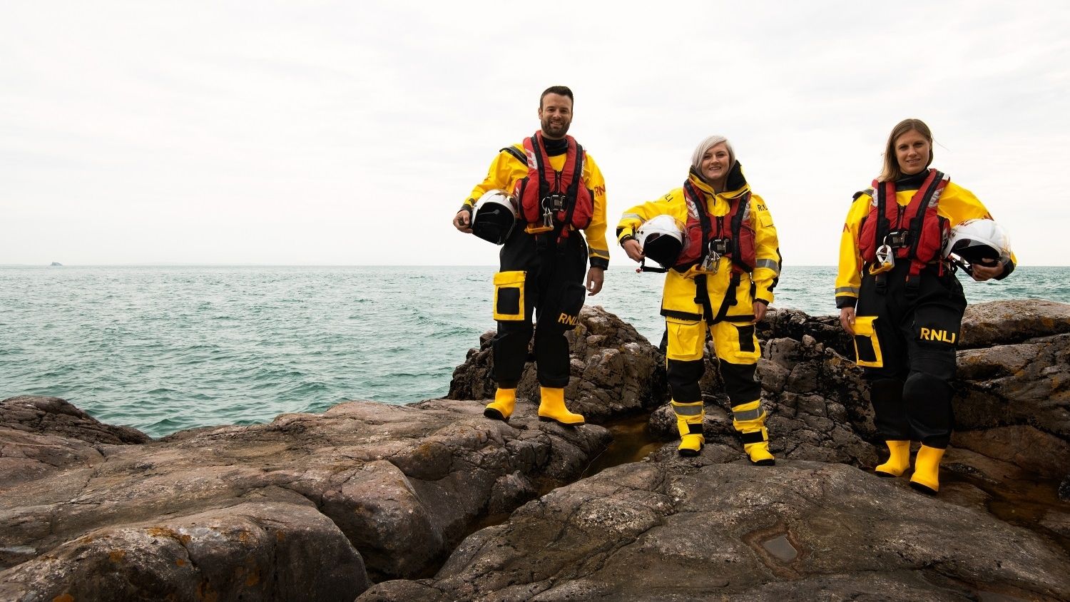 Three crew members are stood on a rock next to the sea smiling and wearing full crew kit