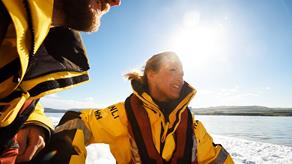 A female crew member onboard a lifeboat at sea wearing RNLI crew kit. She is joined by a fellow male volunteer and they are looking over the horizon together on a sunny day with blue sky.