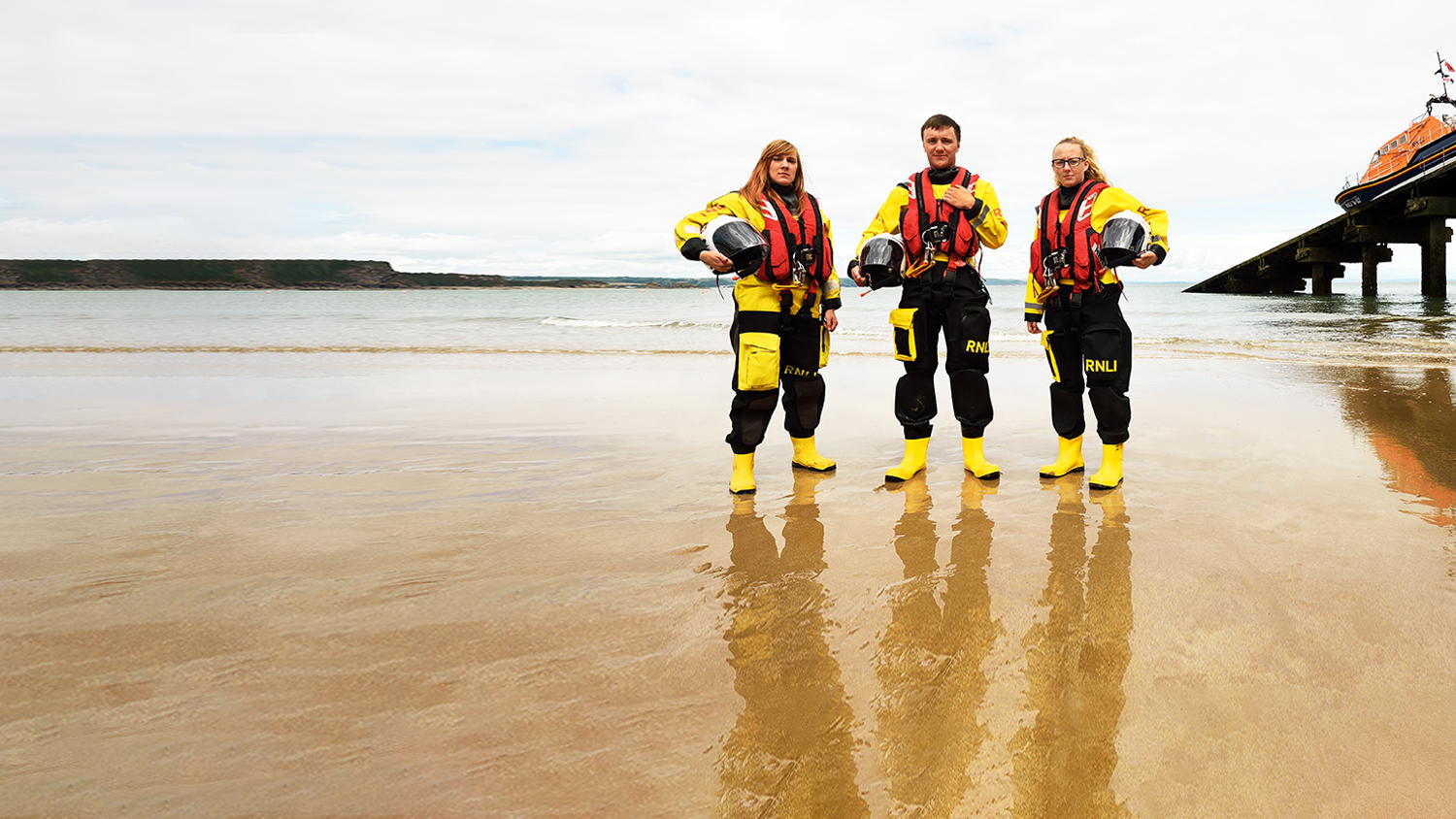Three crew members standing on the sand next to the Tenby Lifeboat Station slipway.