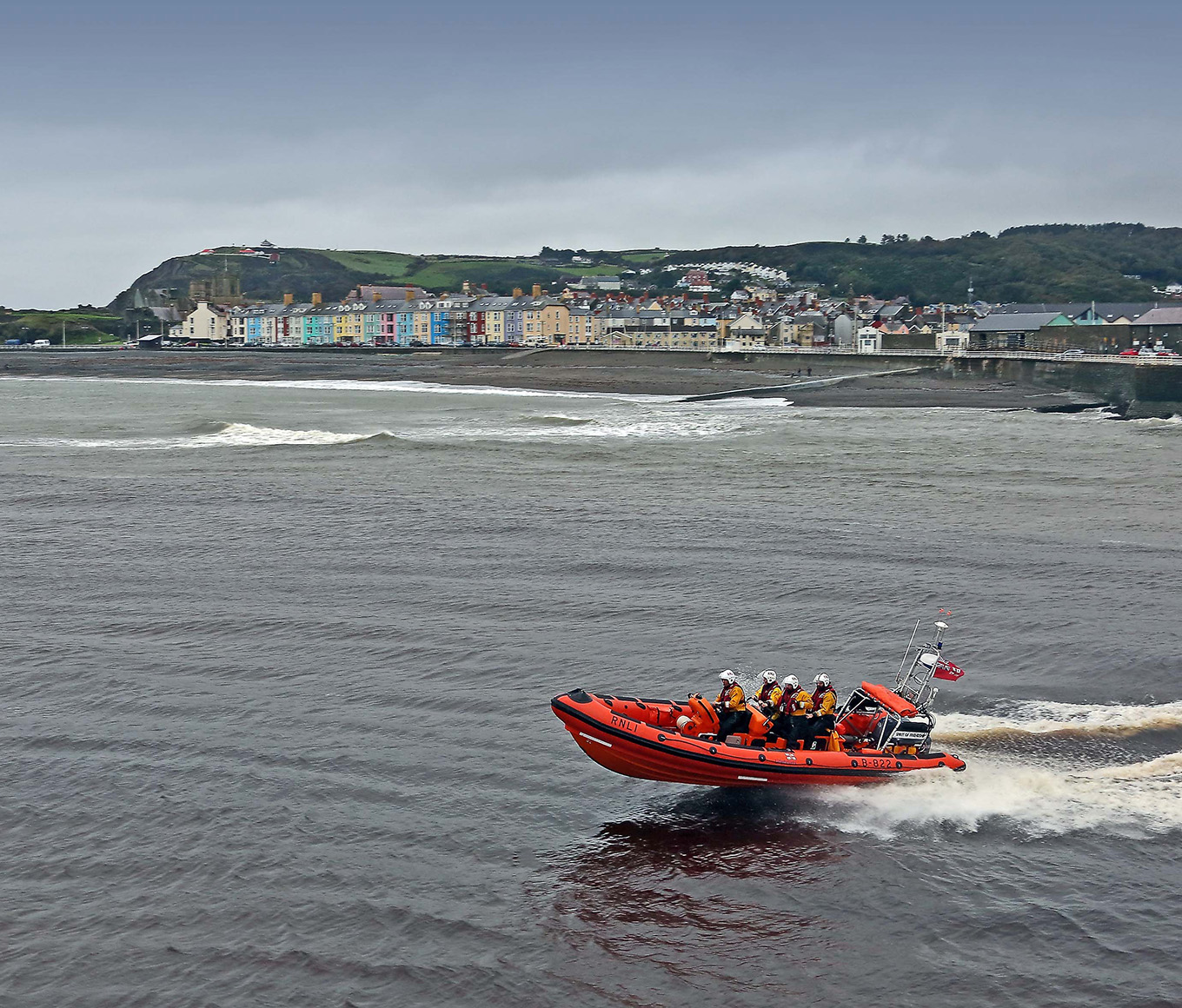 A B class lifeboat with four lifeboat crew members onboard is at sea on an overcast day, with houses and land in the background