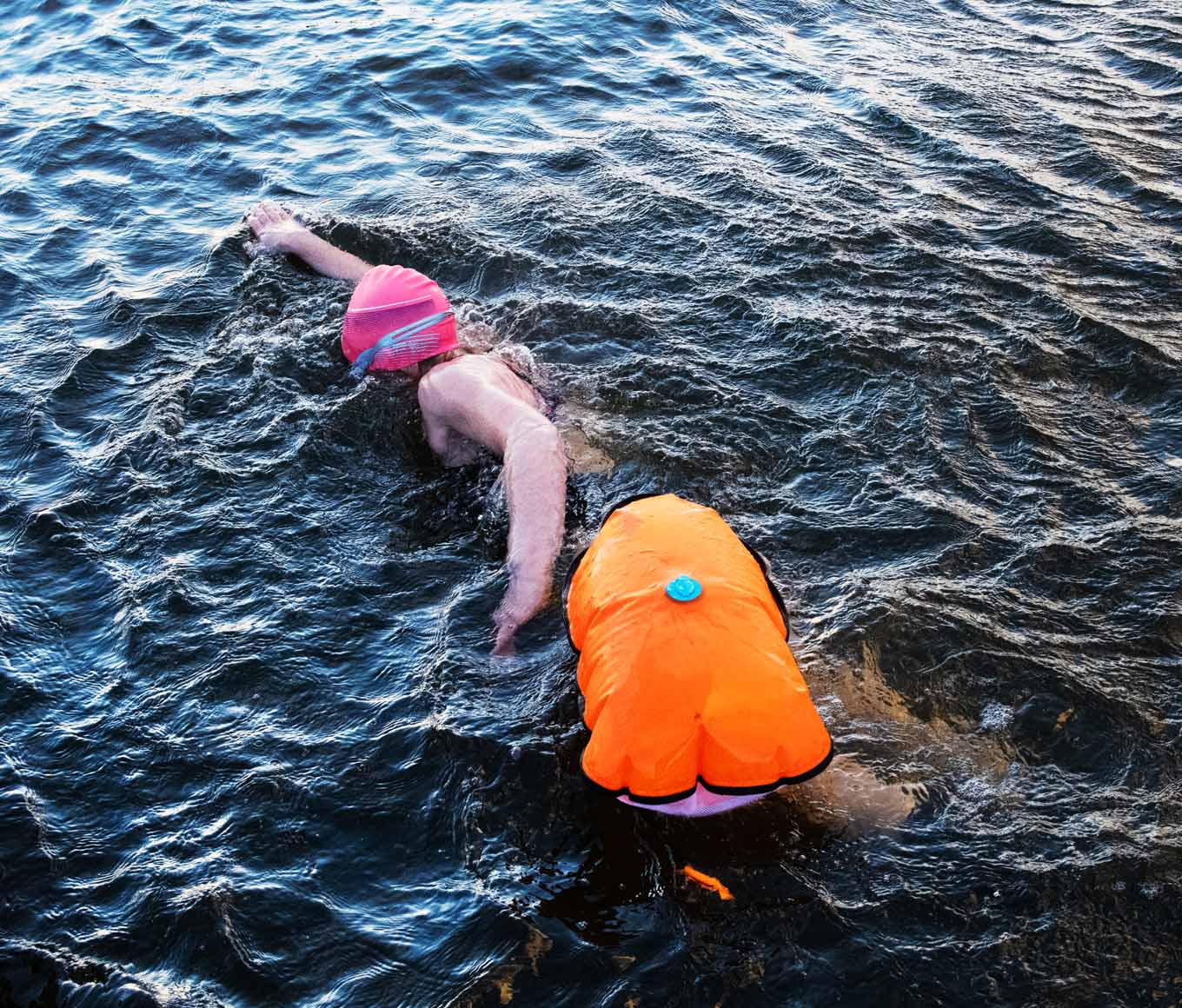 A female cold water swimmer wearing a pink swim cap pulls an orange tow float as she swims through freezing cold seas