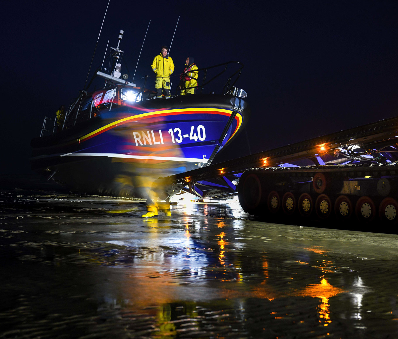 Shannon class lifeboat with two crew on deck is pulled by a tractor along a beach at night