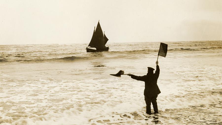 Boulmer pulling and sailing boat Arthur R. Dawes is in the distance at sea. A man is in the shallow water, up to his knees and giving the sailing boat the letter 'J' signal by flags.