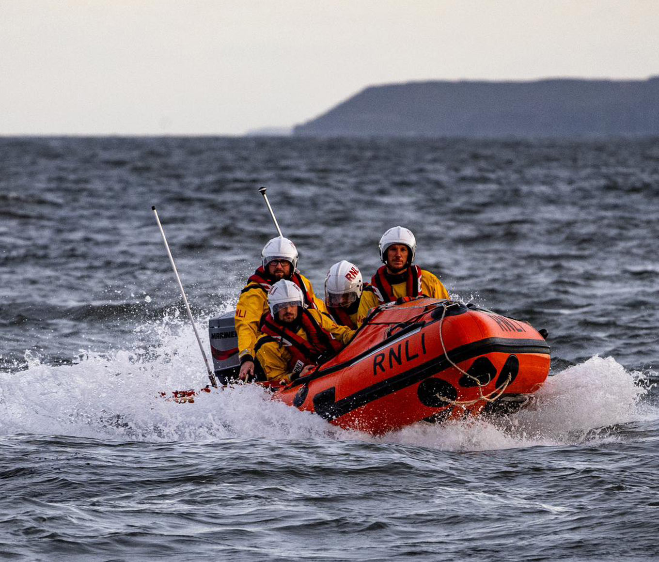 An orange inshore D class lifeboat with four lifeboat crew members onboard powers through calm seas