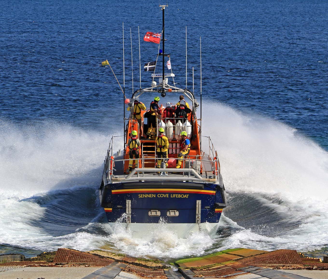 Sennen Cove’s Tamar class lifeboat launches into the waves from the slipway with lifeboat crew in all-weather kit onboard the flying bridge and rear deck