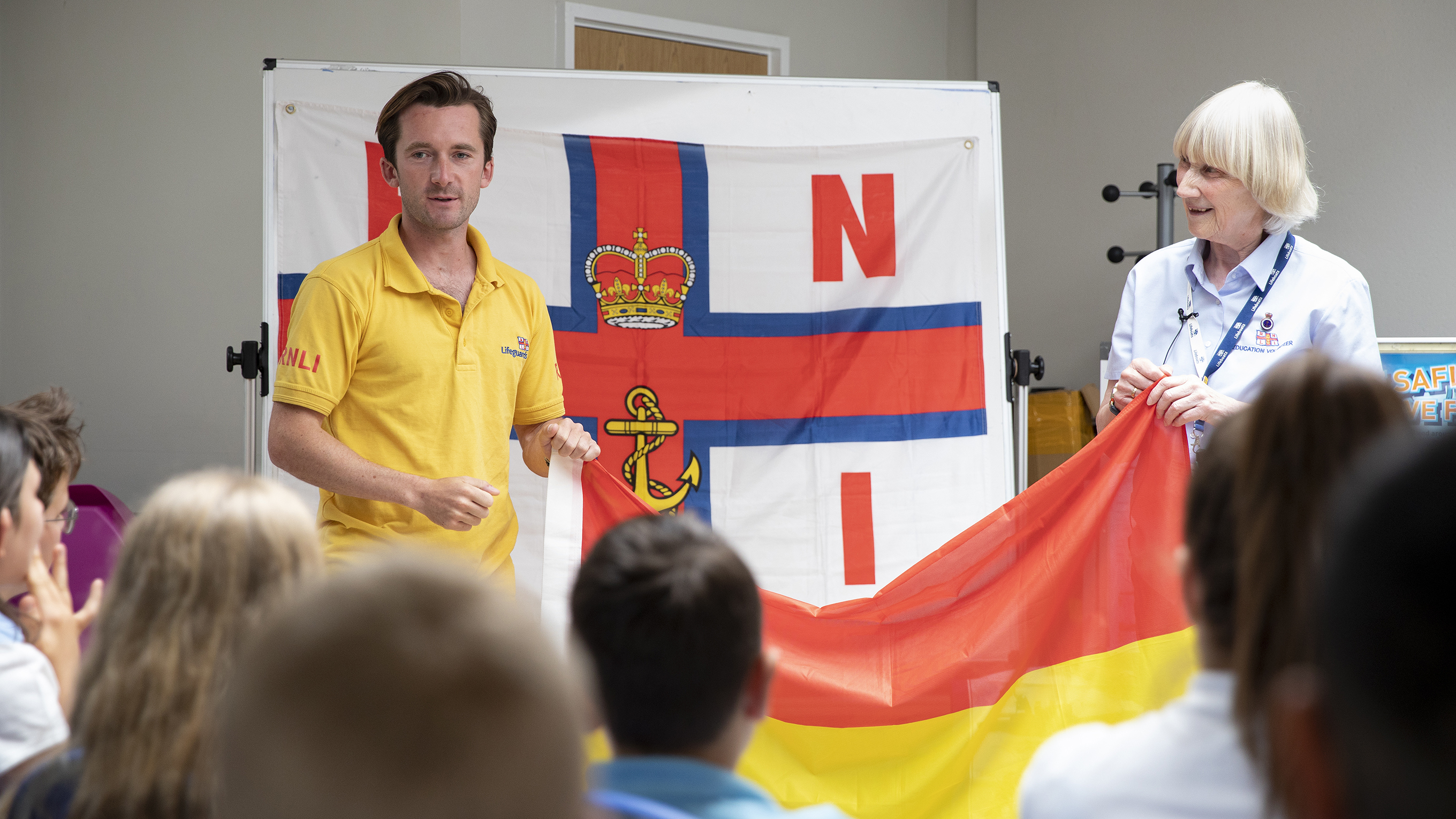 An RNLI lifeguard and a water safety volunteer raise a red and yellow beach flag as they conduct a session in a school classroom.