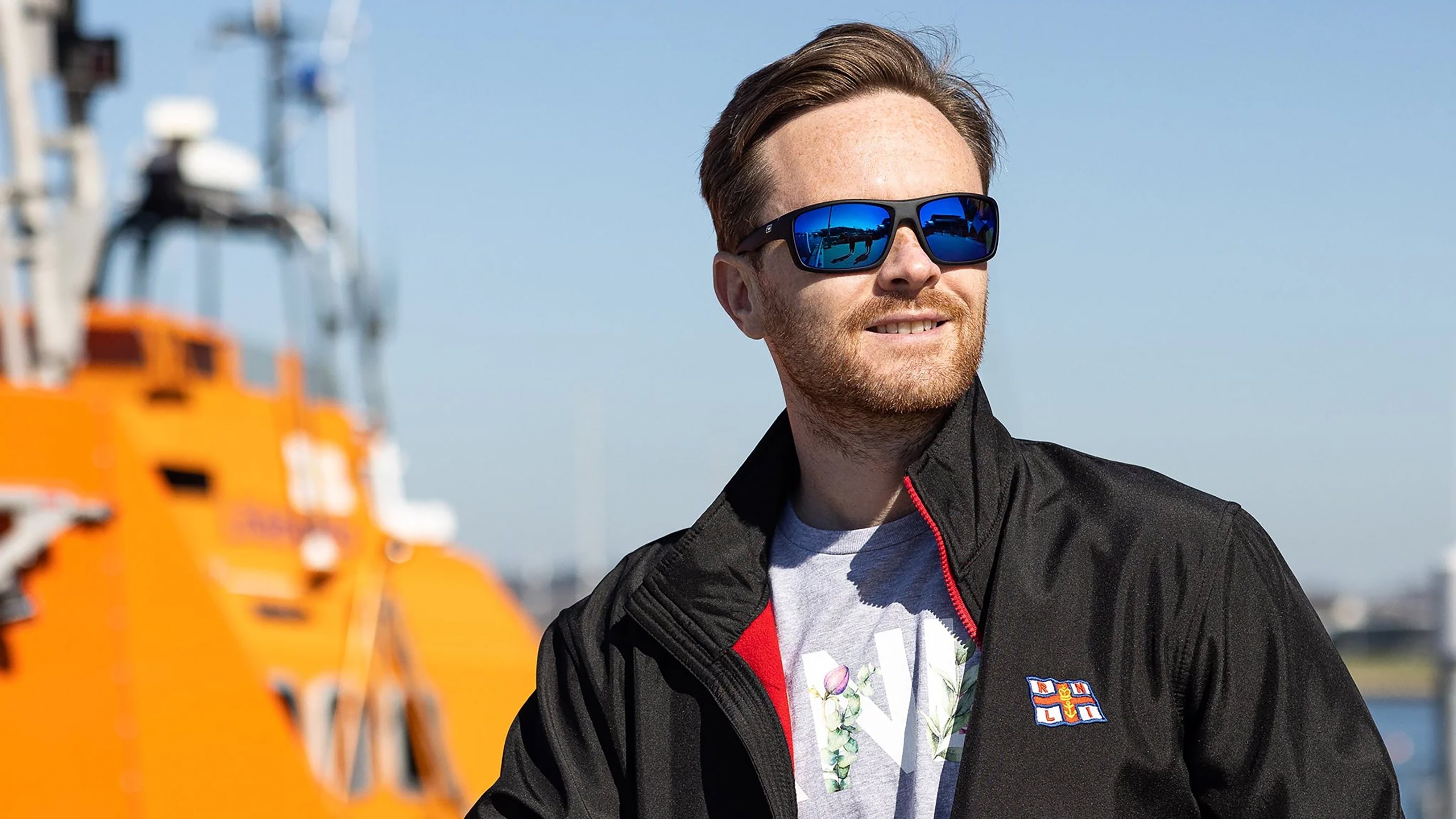 A man standing in front of an orange lifeboat, wearing an RNLI jacket, and a pair of black sunglasses with blue polarised lenses from Dirty Dog