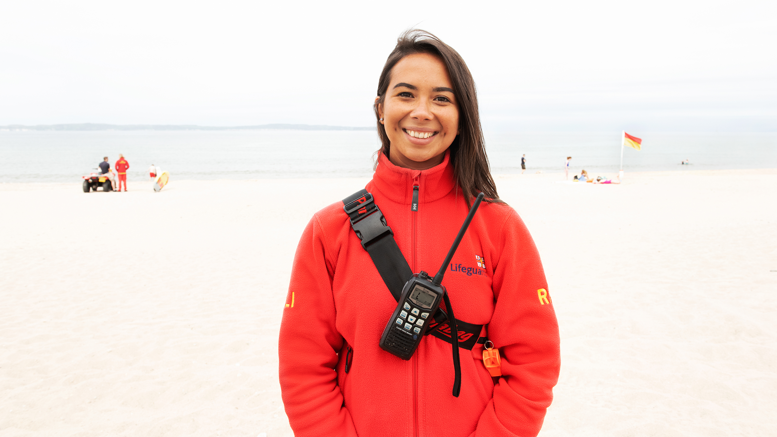 Portush Lifeguard Annie Jagoe, standing on a beach and smiling at the camera. She's wearing a red RNLI lifeguard fleece with a walkie talkie strapped across her. You can see a red and yellow lifeguard flag in the background. 