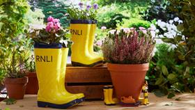 Two yellow welly-shaped planters with flowers sprouting from the tops, on a patio, next to plant pots and small models of a lifeboat, a crew member and a pair of yellow wellies