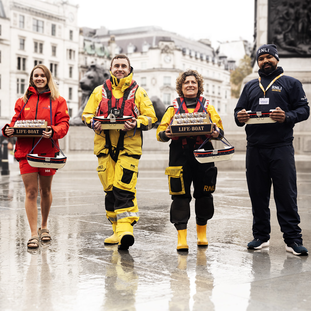 Four RNLI lifesavers stand in a wet Trafalgar Square. From left to right: An RNLI lifeguard, all-weather lifeboat crew member, inshore lifeboat crew member, and a face-to-face fundraiser.