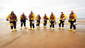 Crew stand on the beach in crew kit holding their helmets