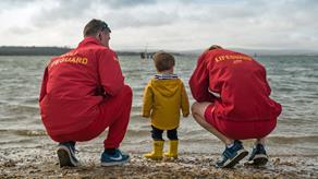 Two RNLI lifeguards crouching down to a young Haven guest on the beach facing the sea with their backs to the camera