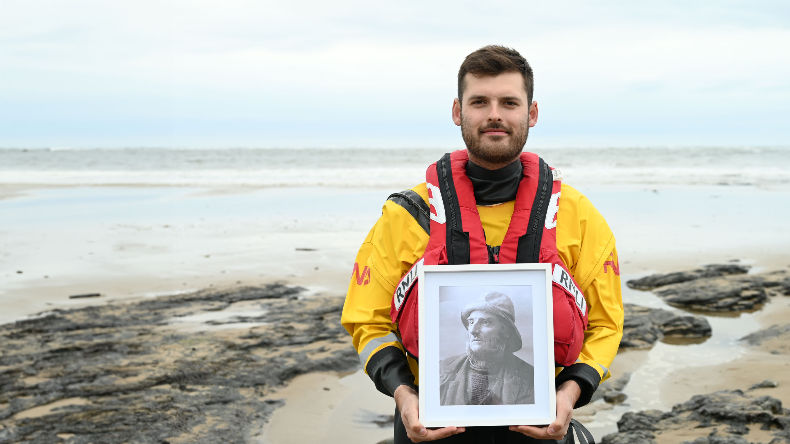 Volunteer Crew Member Luke Picknett is standing on a beach, wearing full lifeboat crew kit. He is holding a framed black and white photograph of his great-great-great grandfather, Coxswain Thomas Picknett.