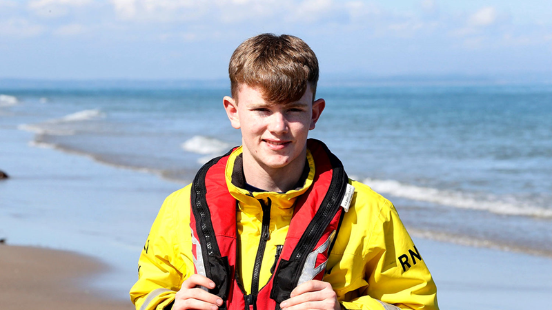 A young male RNLI crew member stands on a beach wearing a yellow RNLI jacket and red lifejacket, he is looking directly into the camera and smiling.