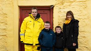 A male crew member in his crew kit is stood outside a yellow building with his two young boys and a woman