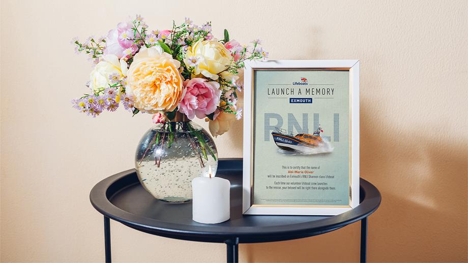 A framed Launch a Memory certificate displayed on a tidy side table along side a candle and vase of colourful blooming flowers
