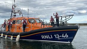 Wells next to Sea's Shannon class lifeboat moving through water with fenders deployed and RNLI people aboard, waving to the camera