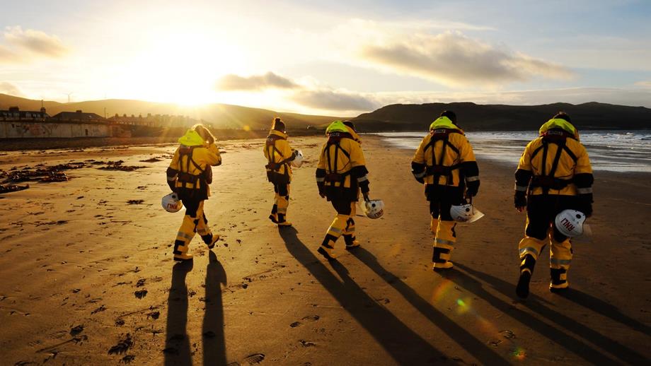 Five lifeboat crew members in RNLI kit, helmets in hand, walk side by side across a Girvan Beach, as the sun sets