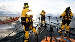 Three lifeboat crew members are stood on the bow of a Shannon class lifeboat at sea, with a B class lifeboat alongside