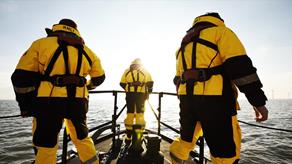 The backs of three Skegness RNLI crew members as they look out to sea from the bow of their Shannon class all-weather lifeboat on a bright sunny day