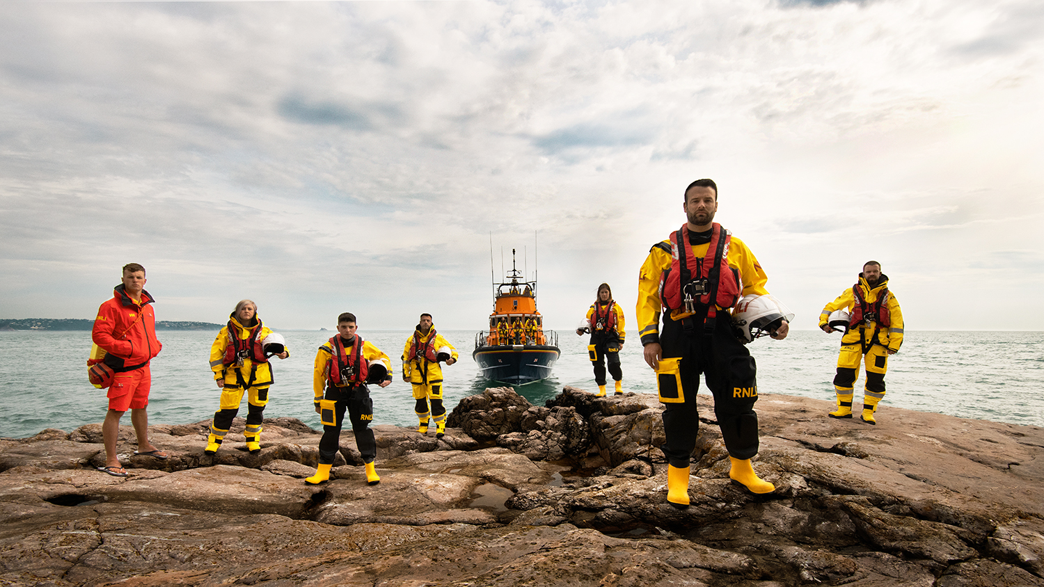 A group of RNLI lifesavers standing on a rock, wearing their kit, with an all-weather lifeboat behind them