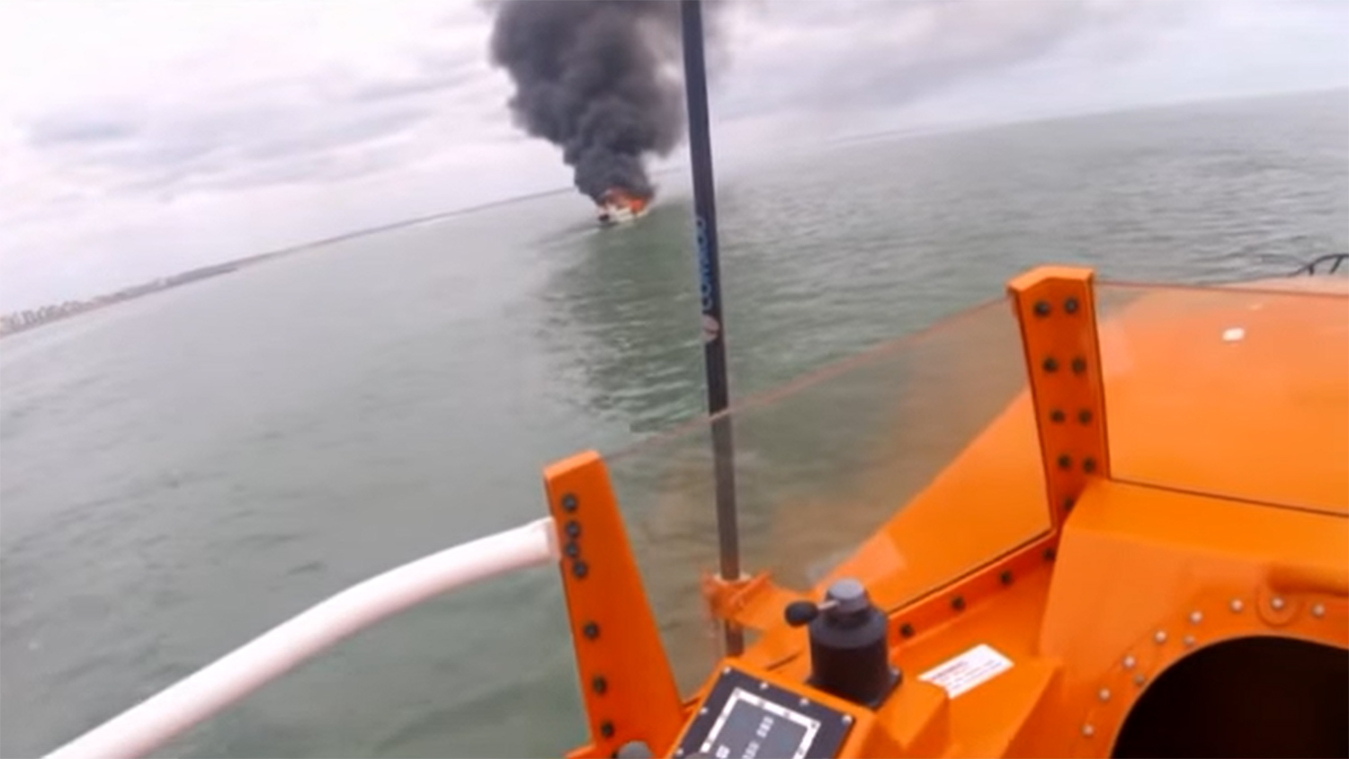 A motor cruiser is on fire, with a large plume of grey smoke billowing into the sky, seen from the vantage point of a lifeboat 