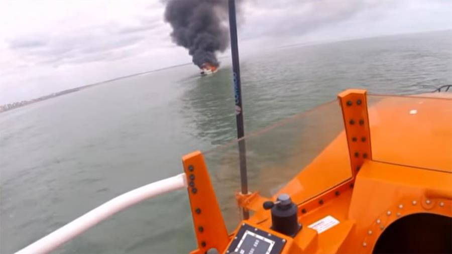 A motor cruiser is on fire, with a large plume of grey smoke billowing into the sky, seen from the vantage point of a lifeboat
