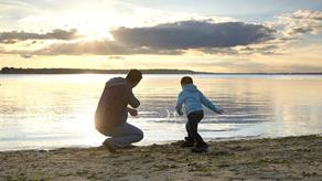 A young child skims stones with an adult on a sandy beach at sunset