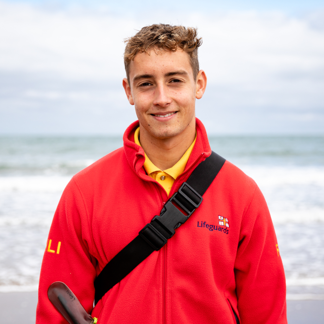 RNLI Lifeguard Hugo is smiling at the camera and is stood in front of the sea on Llangrannog Beach, Wales