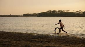 A young boy plays with a wheel in front of a body of water at dusk in rural Bangladesh