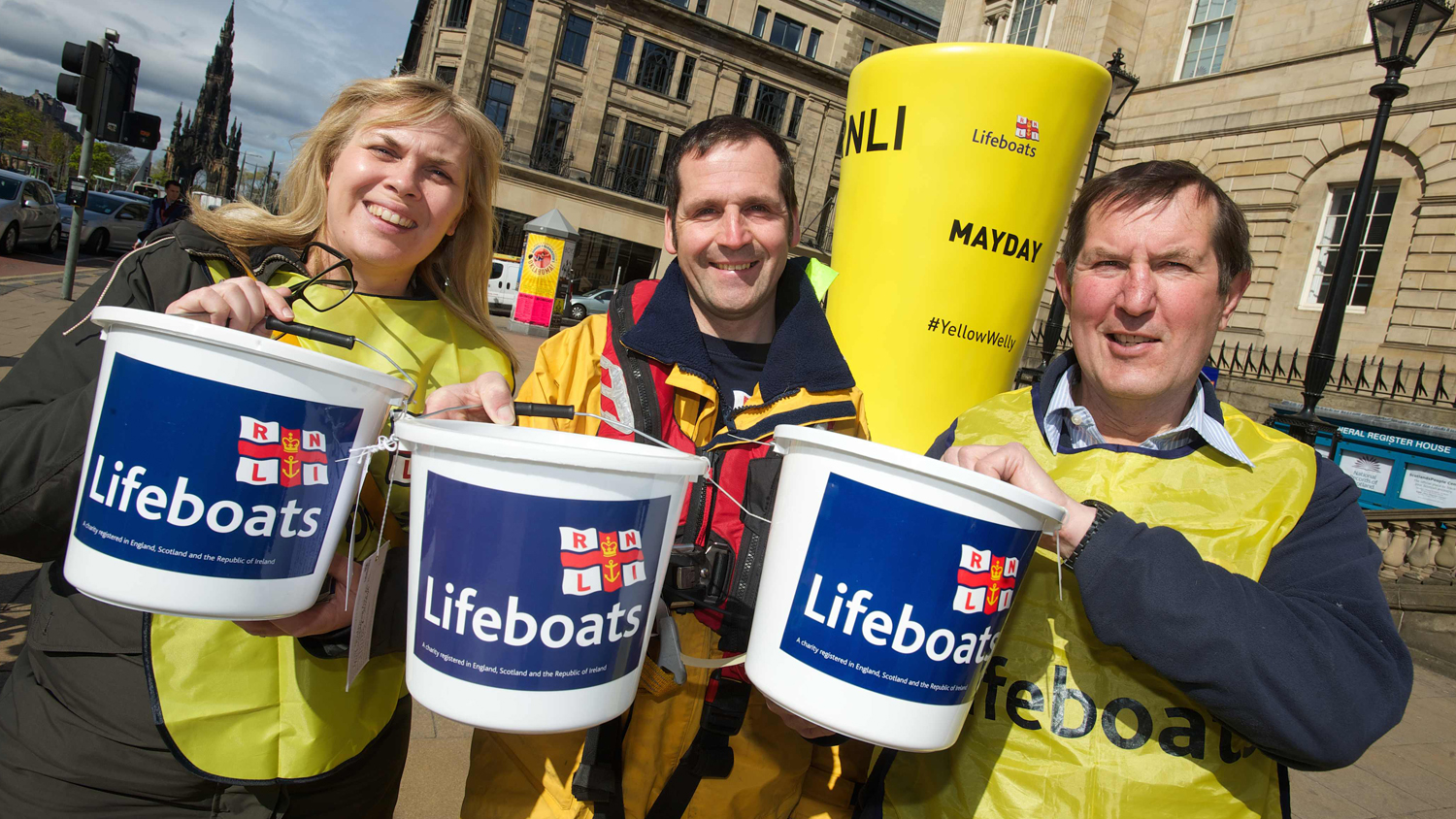 Three RNLI fundraisers standing in front of a giant yellow welly sculpture in Edinburgh. The fundraisers are holding out RNLI buckets used to collect money. 