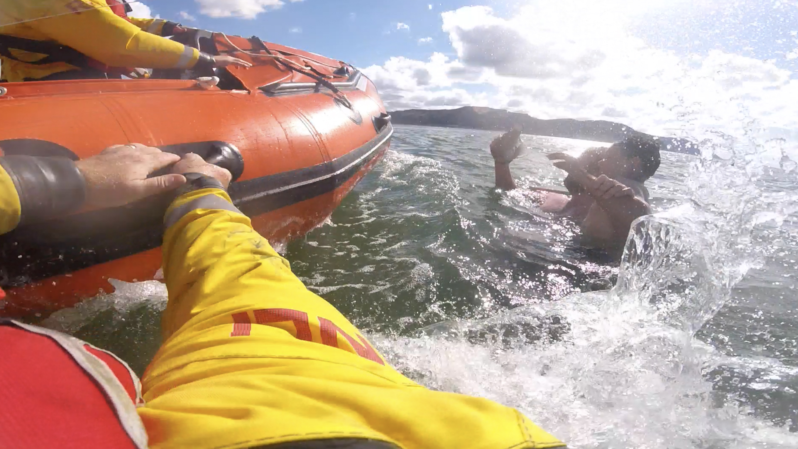 A screenshot from an RNLI lifeboat crew member’s helmet camera footage of a rescue. The crew member is in the water next to the lifeboat. A man is holding a child and they are in deep water, waving their arms.